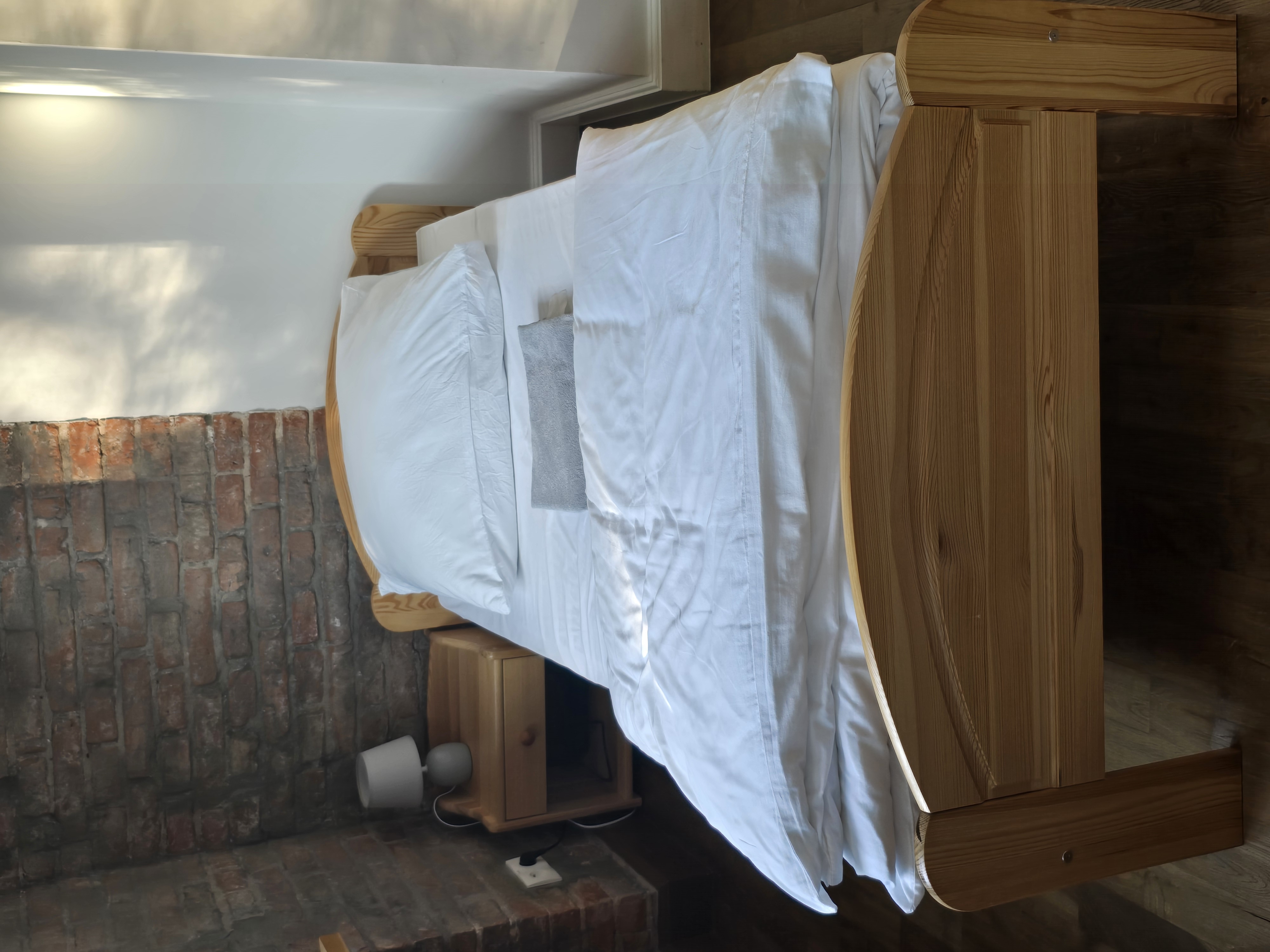 Wygodne świeżo posłane łóżko na tle białej oraz murowanej ściany w jednym z dostępnych pokoi